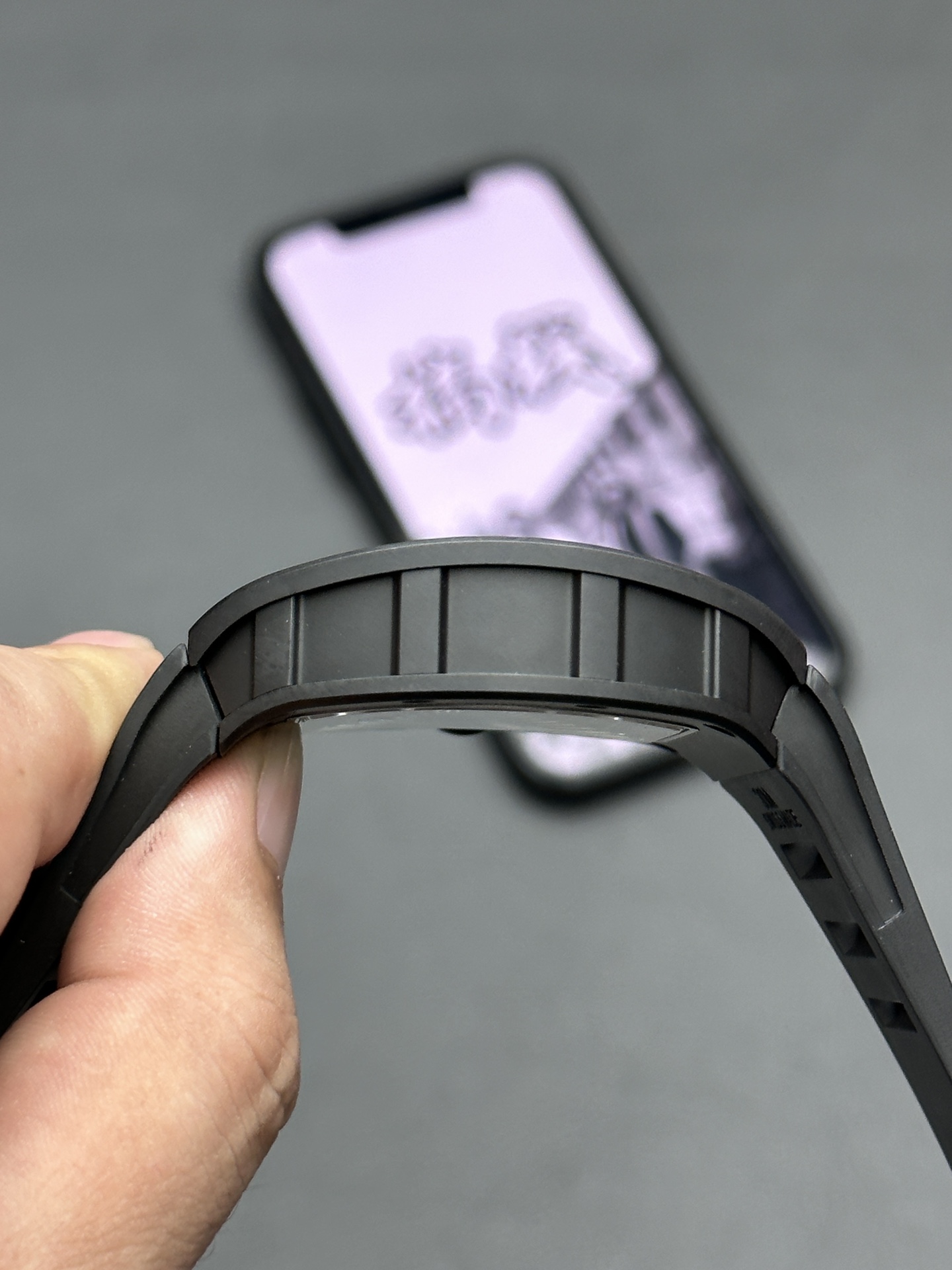 4 - Replica Rolex|Fake Rolex|the best rolex watch factory Close-up of a hand holding a black camera lens or ring, with a blurred smartphone in the background on a gray surface.