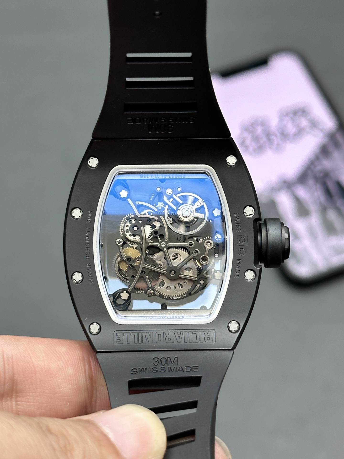 5 - Replica Rolex|Fake Rolex|the best rolex watch factory - Replica Rolex|Fake Rolex|the best rolex watch factory Back view of a Richard Mille skeleton watch showing its intricate gears through a blue-tinted transparent case, with a black rubber strap nearby and 'RICHARD MILLE' engraved on the band edge.