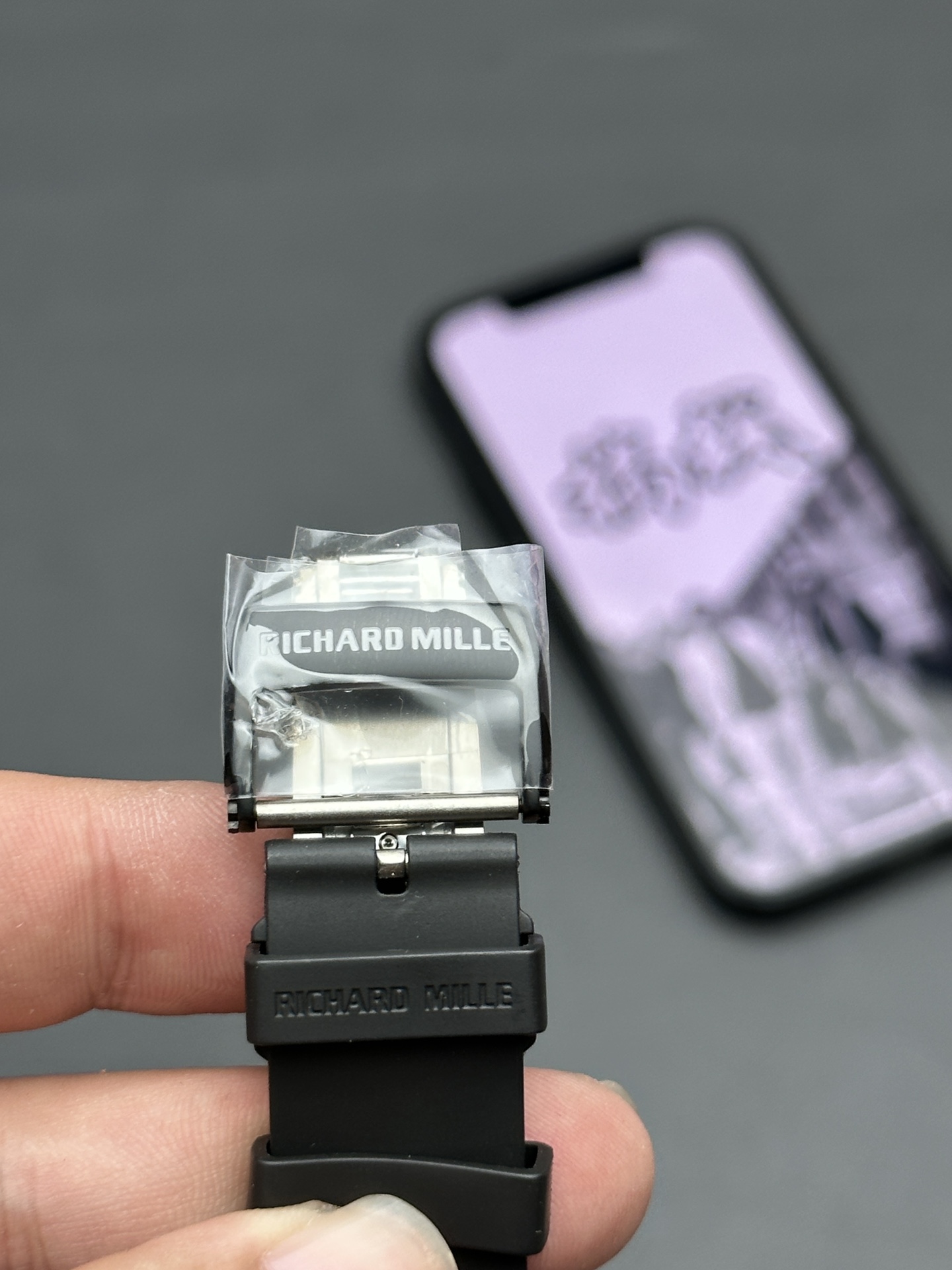 6 - Replica Rolex|Fake Rolex|the best rolex watch factory Close-up of a black Richard Mille watch strap with a clear plastic protector and the brand name visible, held by fingers.